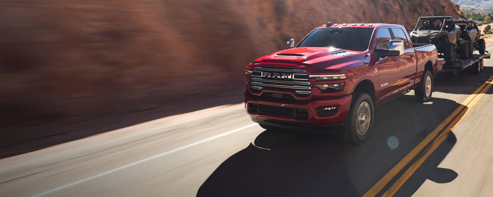 Red 2026 Ram 2500 Crew Cab towing side-by-sides on a trailer along a canyon road; new Ram 2500 trucks for sale at Ross Downing CDJR of Mobile, AL