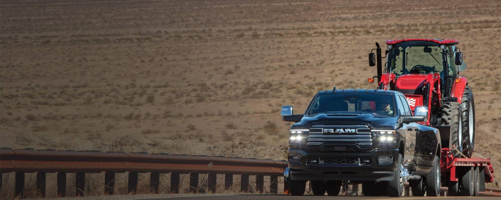 Black 2026 Ram 3500 Crew Cab towing a red tractor on a flatbed trailer through the desert; new Ram 3500 trucks for sale at Ross Downing CDJR of Mobile, AL