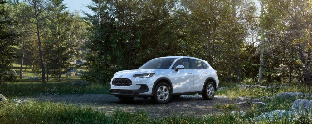 White Honda SUV parked on a gravel path in a wooded area, surrounded by trees and green foliage.
