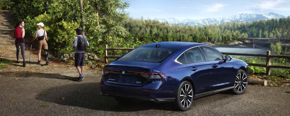 Dark blue Honda sedan parked at a scenic overlook beside a forested lake, viewed from the rear three-quarter angle, with two hikers walking along a trail nearby, surrounded by lush green trees and distant mountains under a clear sky, highlighting a sleek modern car design, outdoor lifestyle, nature travel setting, and premium automotive styling.