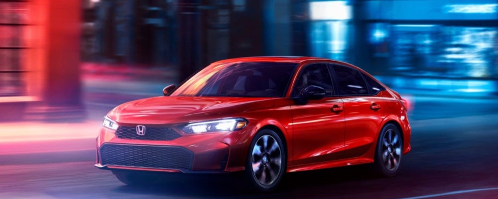 Front-facing red Honda sedan photographed at night in an urban setting, featuring illuminated LED headlights, bold black honeycomb grille with Honda emblem, and glossy red paint reflecting neon city lights, captured in a dramatic low-angle shot that emphasizes sporty performance, modern automotive design, and premium styling against a contemporary city backdrop.