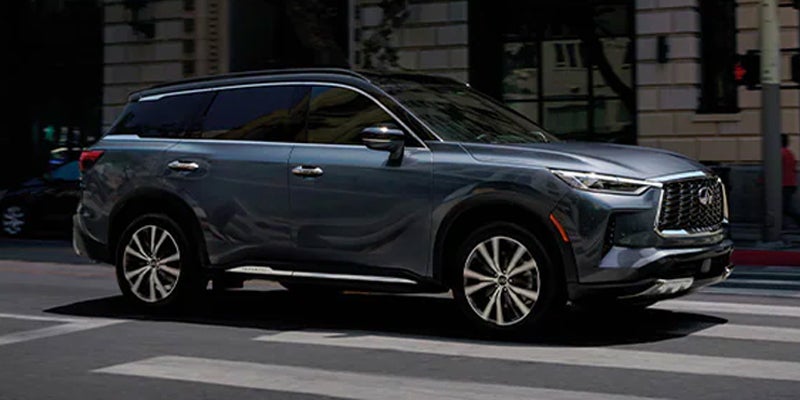A dark grey SUV INFINITI QX60 driving on a city street with buildings in the background.