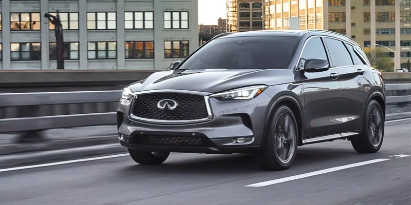 A gray Infiniti QX50 SUV driving on a highway, with city buildings blurred in the background.