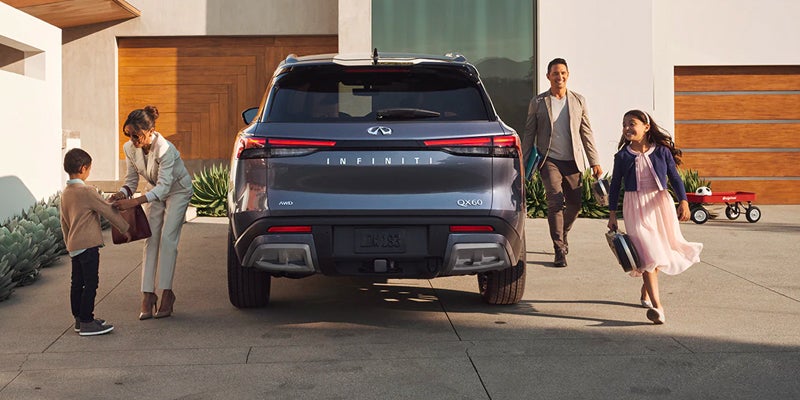 Rear view of a dark gray Infiniti QX60 parked in a driveway. A family of four approaches or moves away from the car.