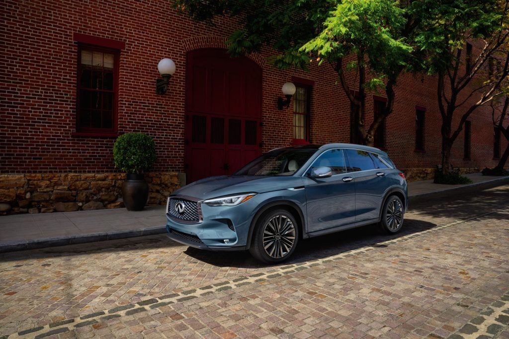 A teal Infiniti QX50 crossover SUV parked on a cobblestone street, next to a red brick building with trees.
