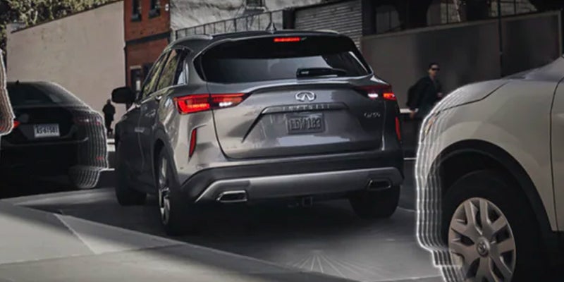 Rear view of a grey SUV Infiniti QX60 with brake lights on, parked between two other cars on a city street.