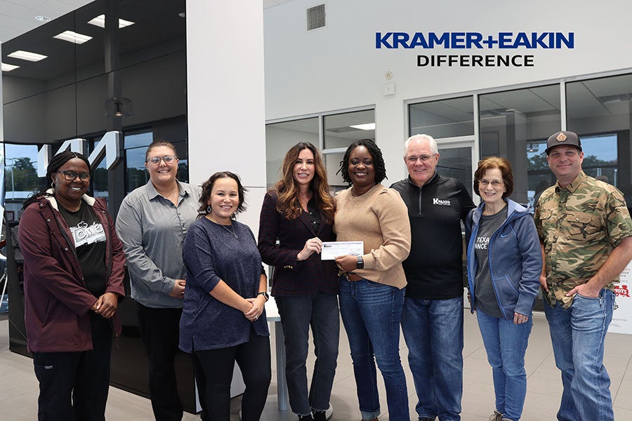 A group of eight people stands smiling indoors, with one woman holding a check. The setting appears professional, with "Kramer + Eakin Difference" on the wall.