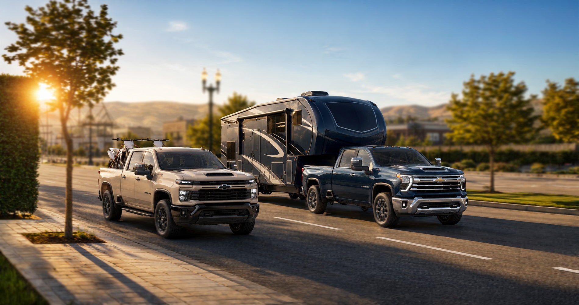 Two 2026 Chevrolet Silverado 2500 trucks—a tan model carrying dirt bikes and a blue model towing a large fifth-wheel RV—driving side by side through a suburban street in Pittsburg, California, with hills and trees in the background under golden evening light, emphasizing the Silverado’s power and capability.
