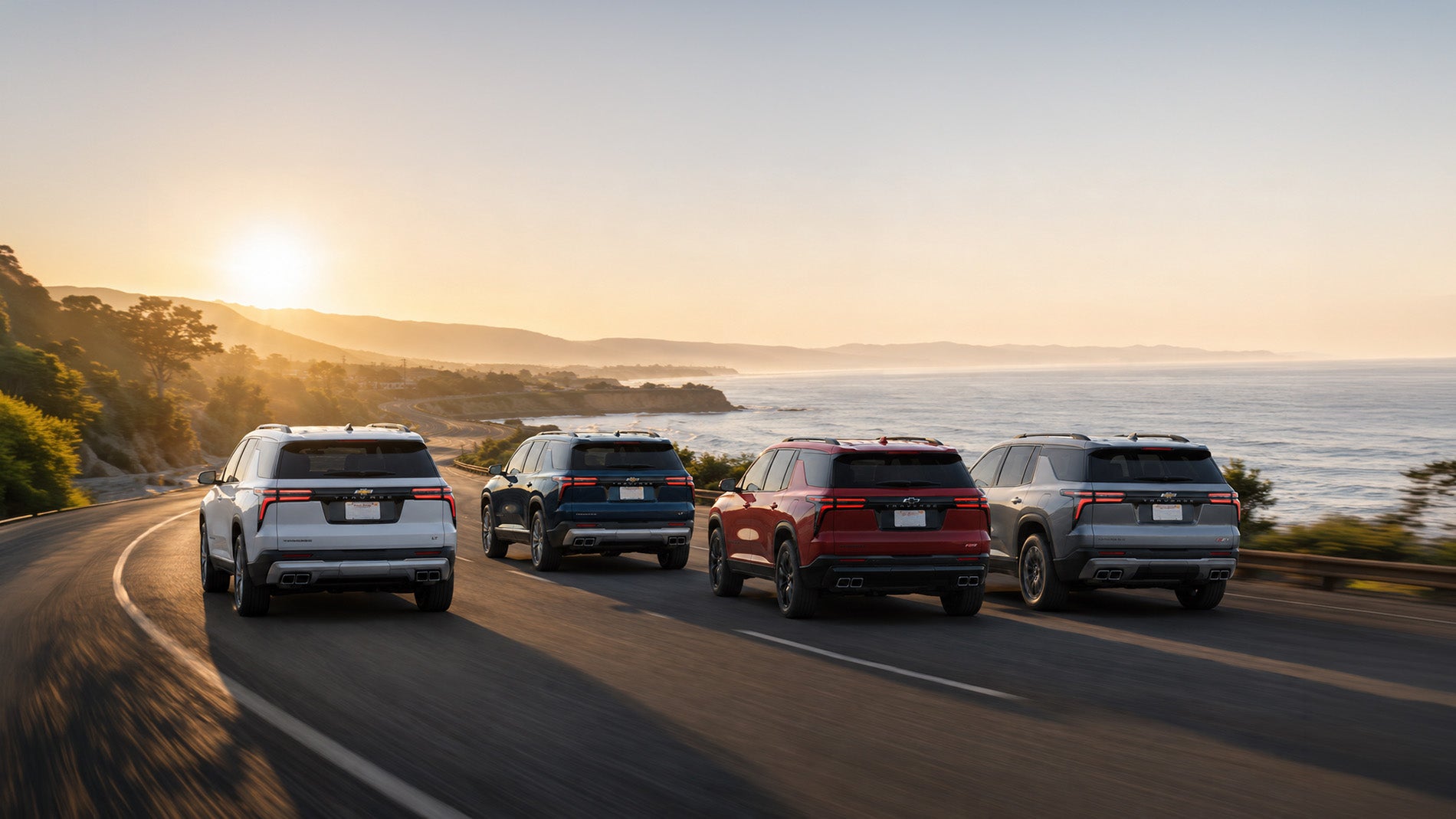 Four 2026 Chevrolet Traverse SUVs driving along a coastal highway at sunset, with the Pacific Ocean and rolling hills in the background, illustrating style and versatility across Traverse trim levels.