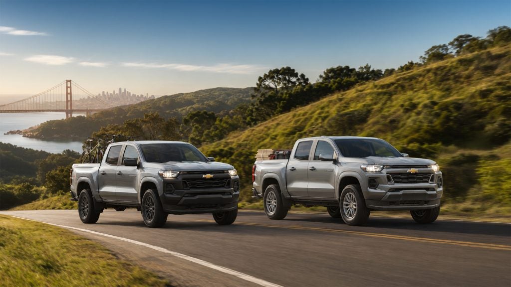 Two silver 2026 Chevrolet Colorado pickup trucks drive side by side along a coastal highway overlooking the Golden Gate Bridge and San Francisco Bay, one carrying bicycles and the other loaded with gear, illustrating the truck’s versatility and trim options available at Delta Chevrolet in Pittsburg, CA.