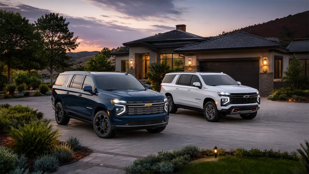 A blue and white 2026 Chevrolet Suburban parked side by side in a modern residential driveway at sunset, highlighting distinct styling between luxury and off‑road Z71 trims available at Delta Chevrolet in Pittsburg, CA.