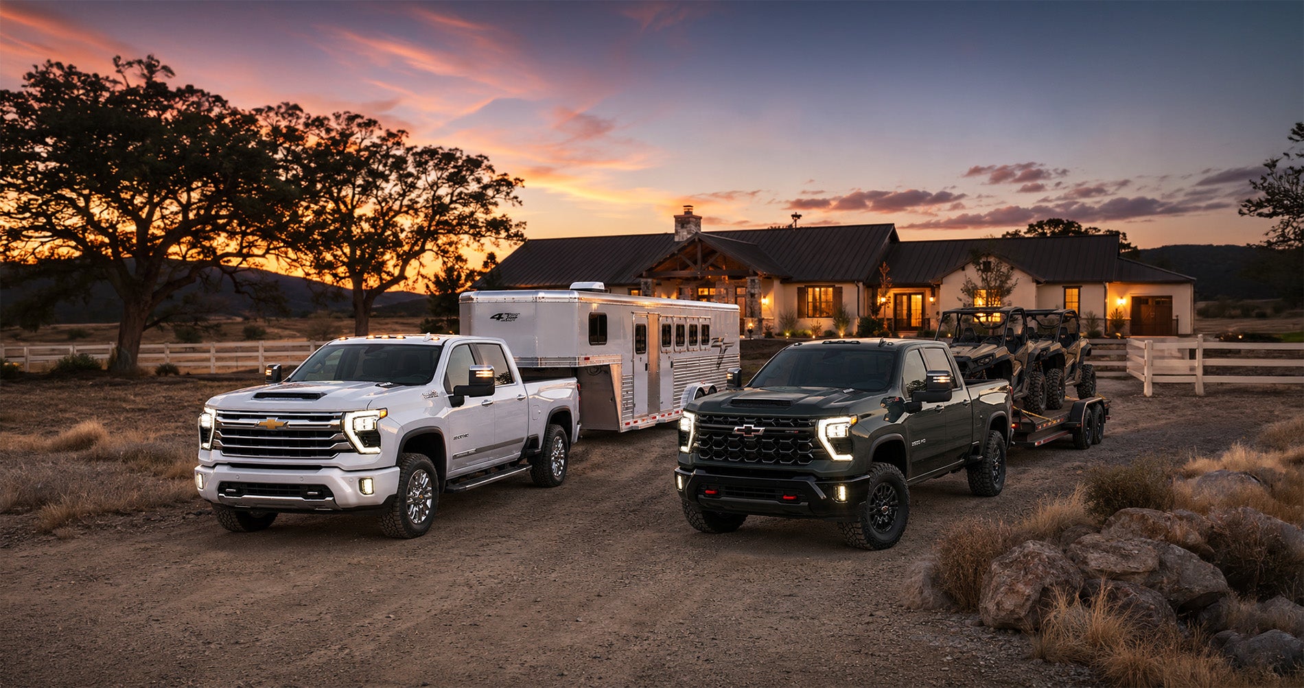 Two 2026 Chevrolet Silverado 2500 trucks—a white High Country towing a livestock trailer and a black Z71 towing off-road vehicles—parked on a ranch driveway at sunset near Pittsburg, California, with warm house lights and trees in the background, highlighting the trucks’ towing strength and versatility.