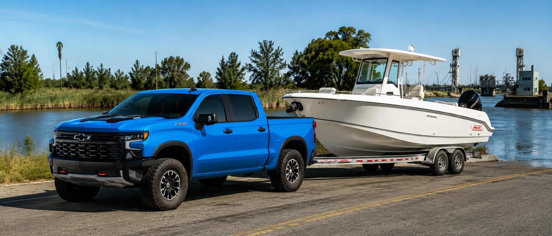 Blue Chevrolet Silverado ZR2 towing a white center-console fishing boat on a tandem-axle trailer beside a marina and boat launch.