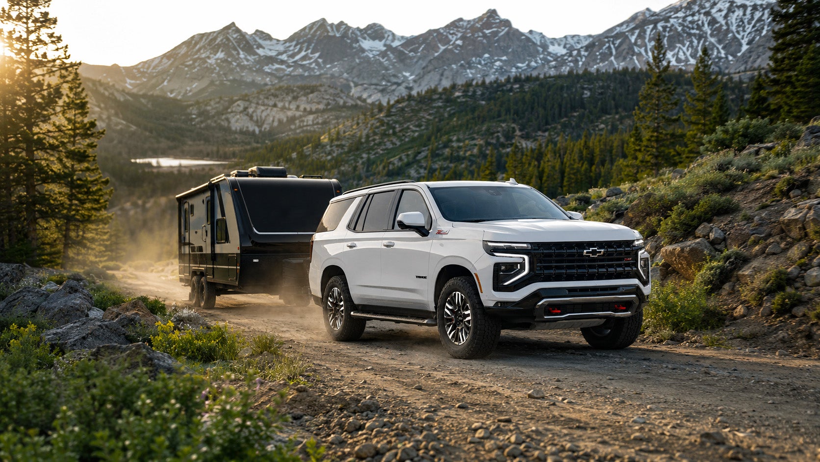 A white 2026 Chevrolet Tahoe Z71 towing a black travel trailer on a mountain road at sunrise, with snow‑capped peaks and evergreen trees in the background, illustrating Tahoe’s power and off‑road performance.