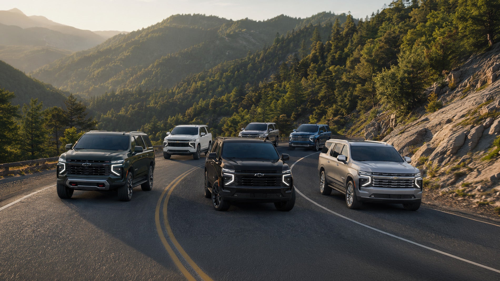 Six 2026 Chevrolet Suburban SUVs in black, white, silver, and blue drive along a winding mountain road surrounded by pine trees and rocky terrain at sunset, showcasing the range of Suburban trim levels available at Delta Chevrolet in Pittsburg, CA.