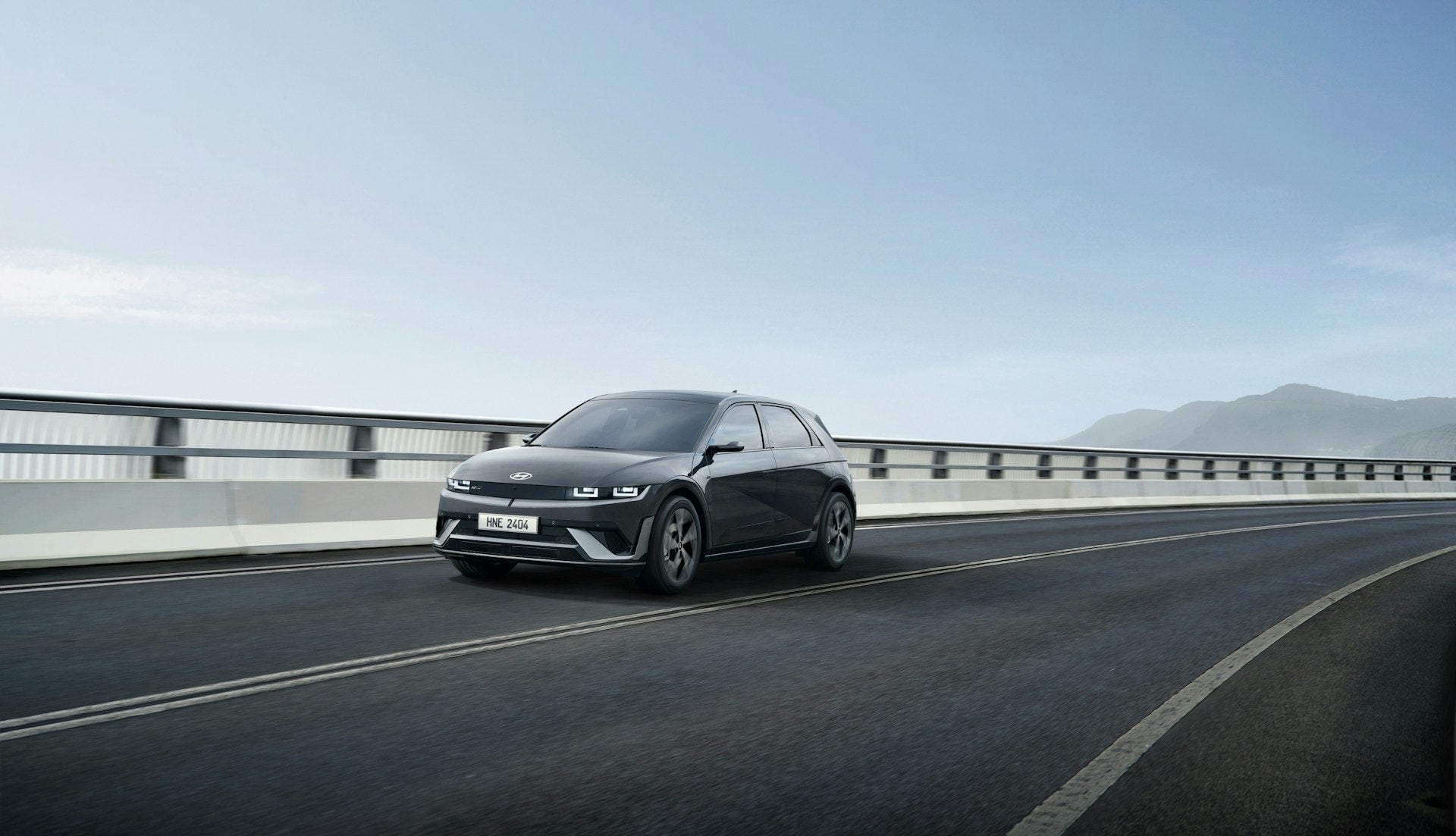 Front view of a gray Hyundai car with its headlights on, driving on a road next to a body of water and mountains.