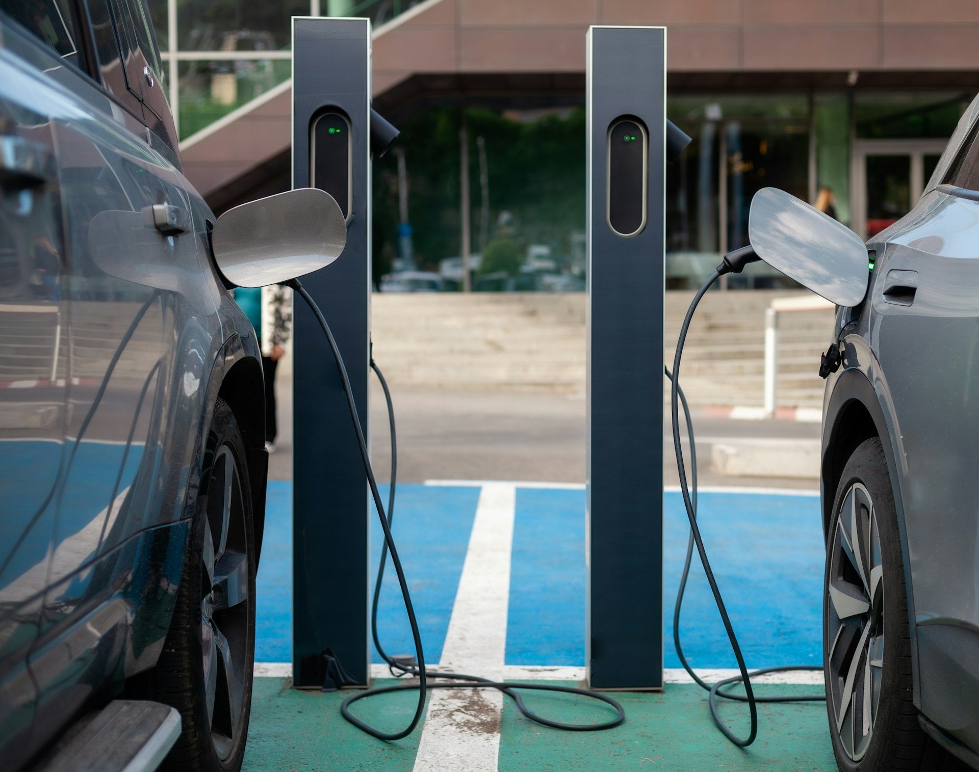 Two charging cable connects a white electric car to a wall-mounted charging station with a green light bar.