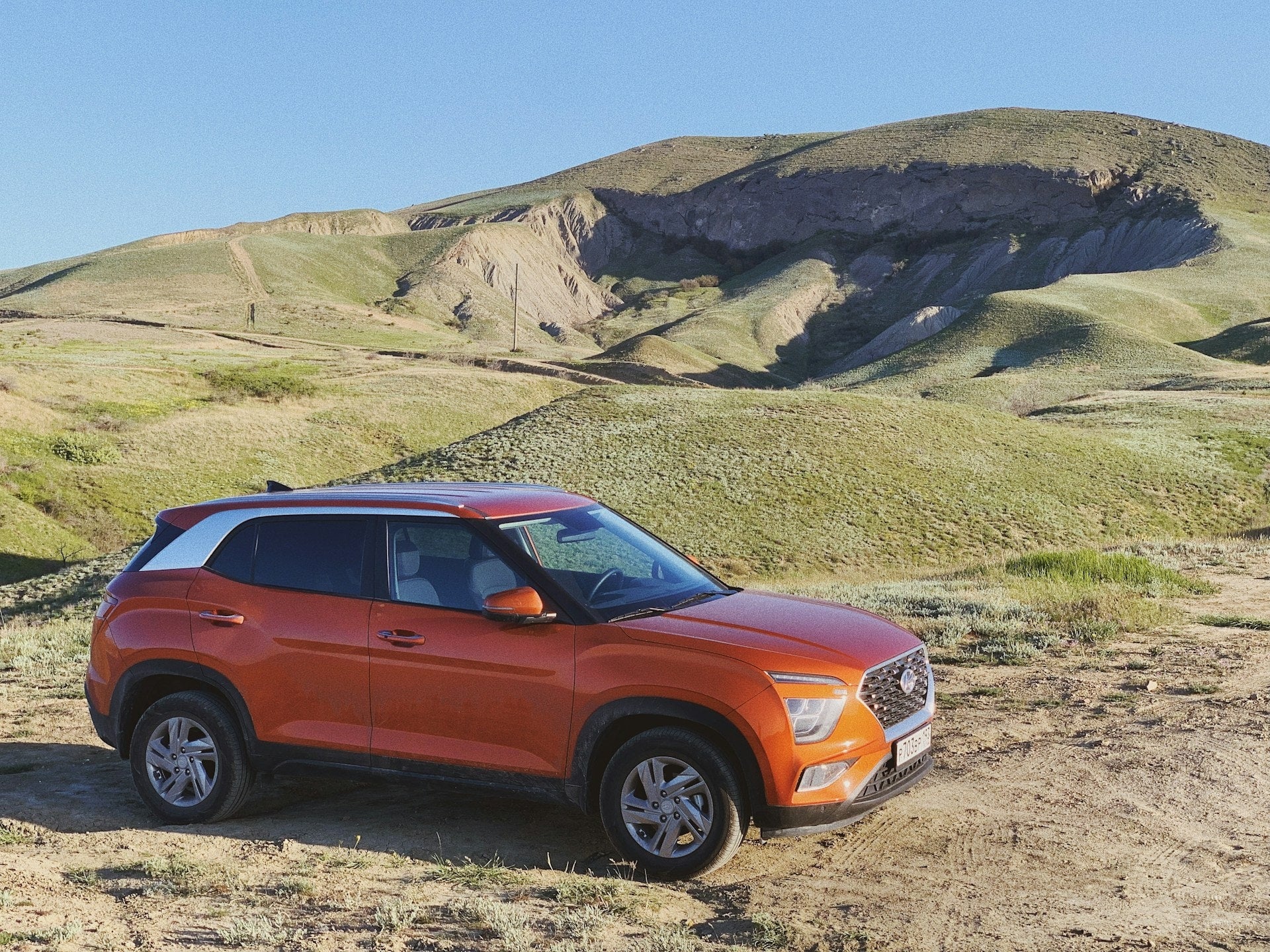 Light orange car parked on a stone driveway, with a couple chatting near the car's front. Modern .