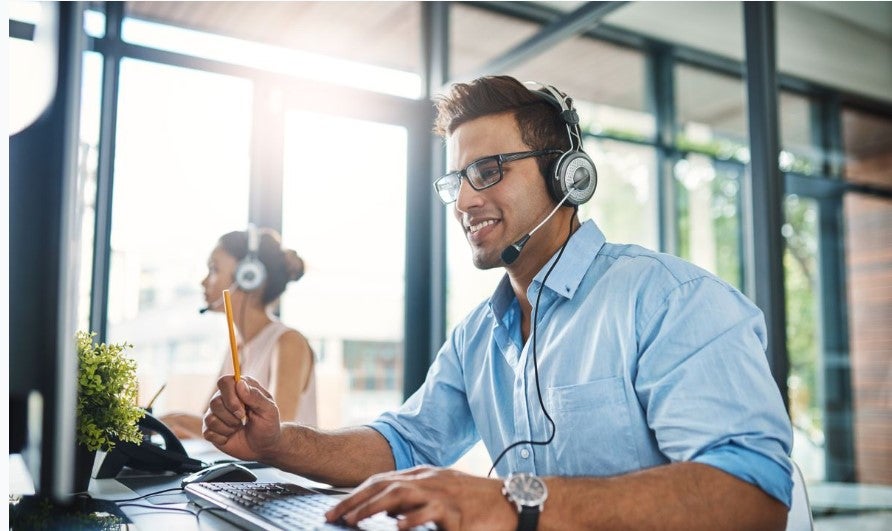 Customer service representative wearing a headset, smiling while working at a desk and taking notes during a phone call in a modern office environment.