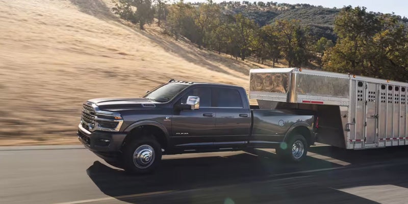 A dark gray Ram truck with a silver trailer attached is driving on a highway, with hills and trees in the background.