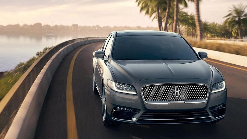 A grey Lincoln sedan drives on a coastal road lined with palm trees.