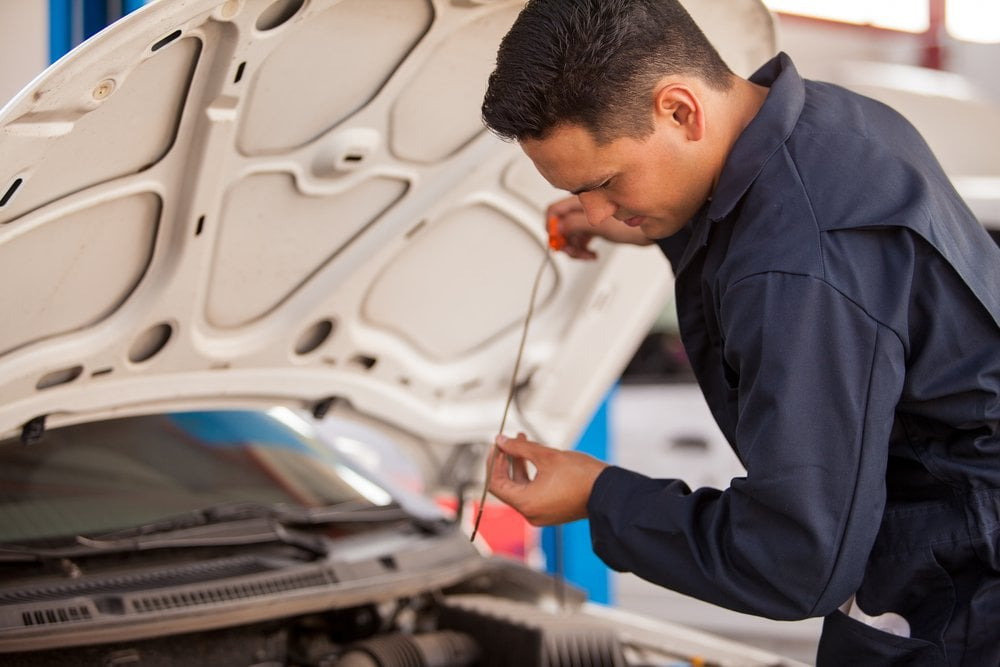A mechanic in blue coveralls checks a car's oil with a dipstick in a garage.
