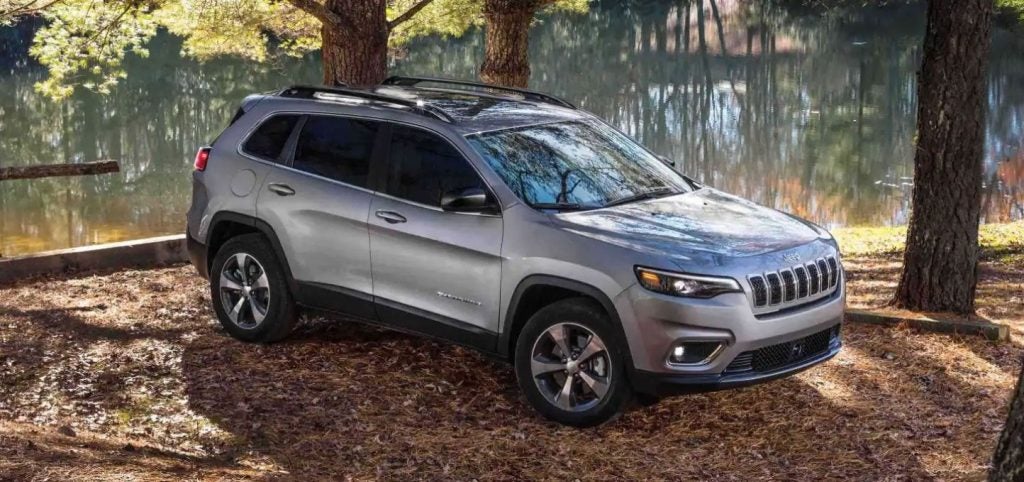 Silver Jeep Cherokee SUV with a roof rack parked on dry leaves by a lake, trees reflected in the water.