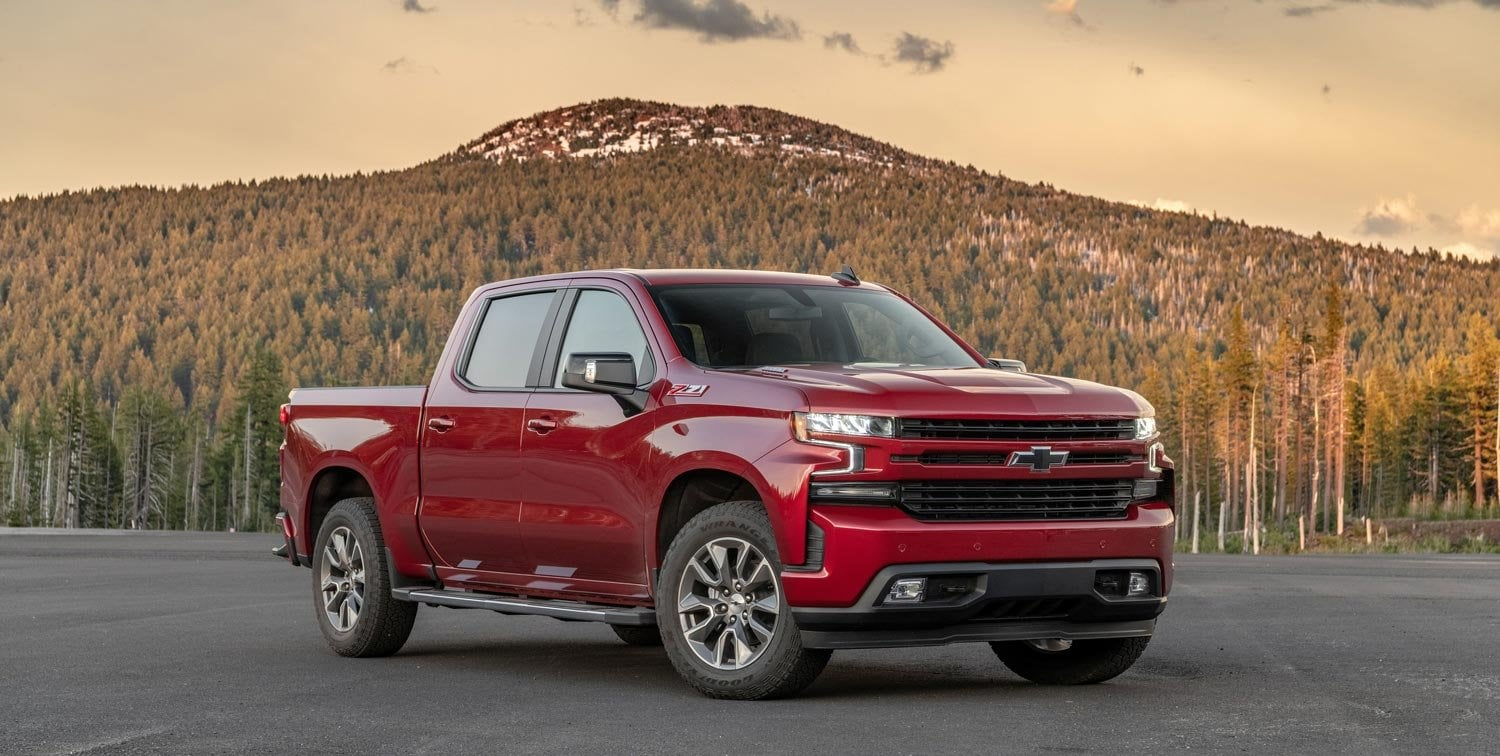 A Red Chevrolet Silverado pickup truck kicks up dust as it drives rapidly over a dirt field during the day.