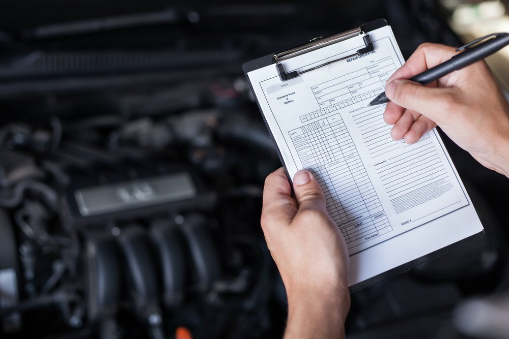 A mechanic in a black shirt writing on a clipboard with a black pen, blurred car engine in the background.