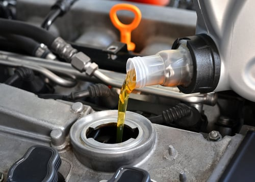 A hand pours golden motor oil from a gray container into a car's engine.