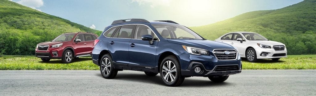 A blue Subaru Outback, red Subaru Forester, and white Subaru Legacy parked on a road with green hills in the background.