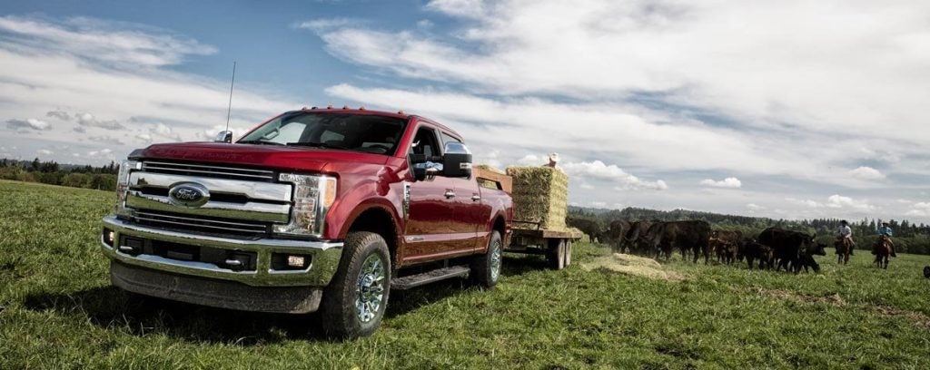 Red Ford pickup truck with a hay trailer in a green pasture with cows and riders in the background.