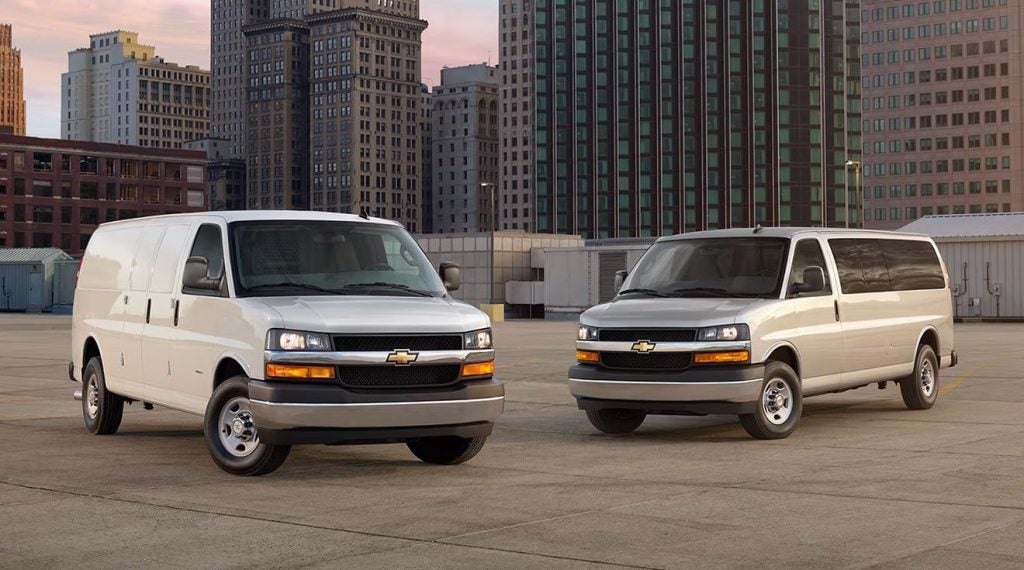 Two Chevy Express vans, a white cargo van and a silver passenger van, parked on an urban rooftop.