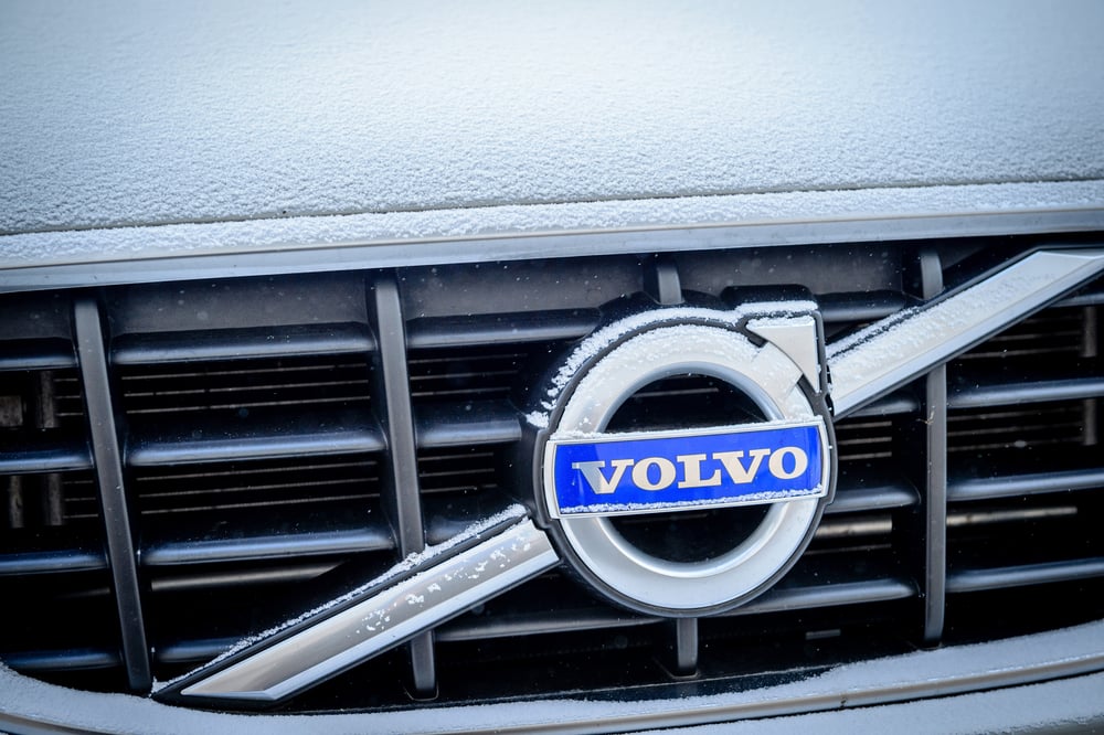 Close-up of a silver Volvo emblem with a blue VOLVO text bar, partially covered in snow on a black car grille.