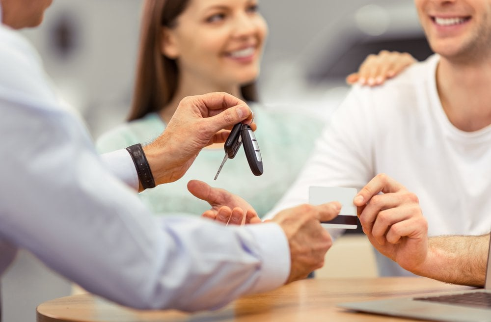 Couple signing paperwork at a dealership while salesman holds up car keys