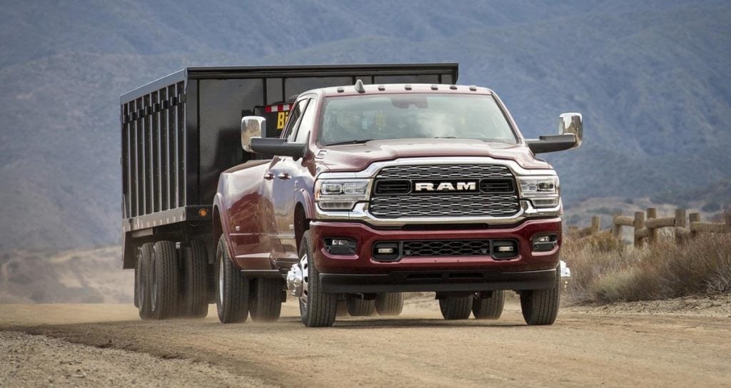 A maroon Ram 3500 heavy-duty pickup truck pulls a black dump trailer down a dirt road with mountains in the background.
