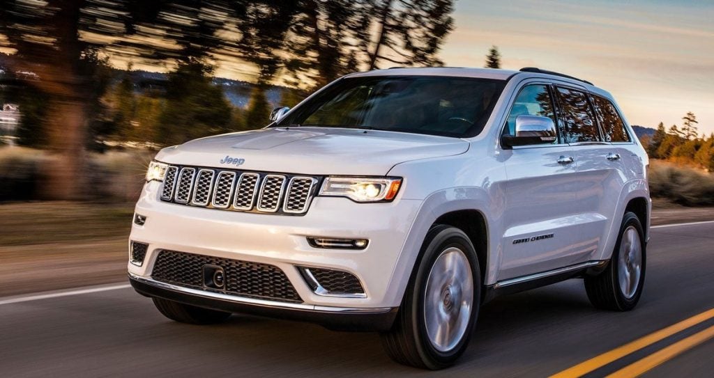 White Jeep Grand Cherokee SUV driving on a road surrounded by trees and mountains at sunset.