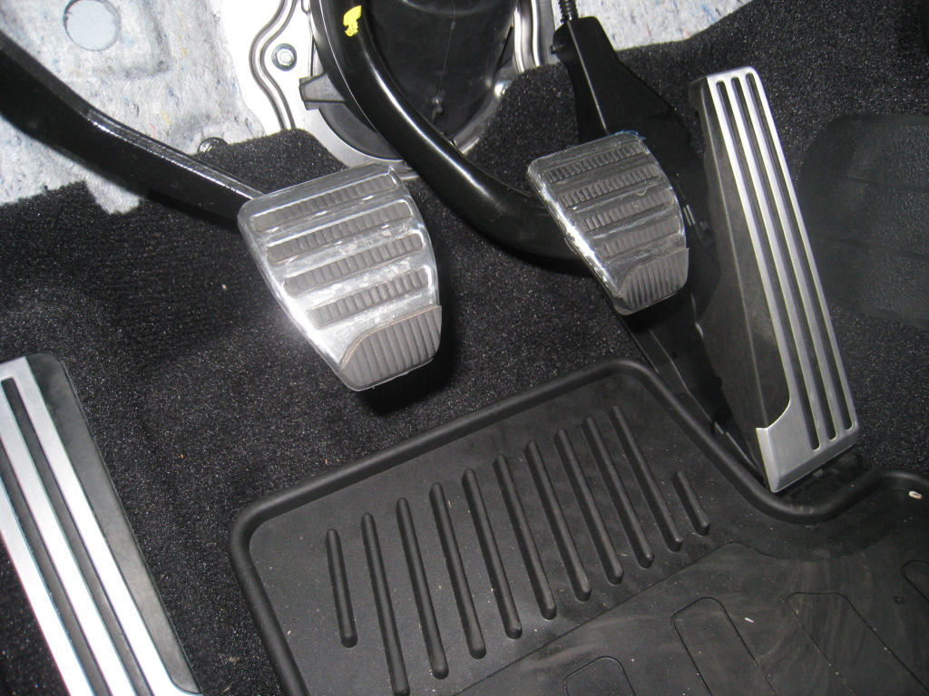 Three car pedals on a dark floor: clutch (left), brake (center), and accelerator (right).