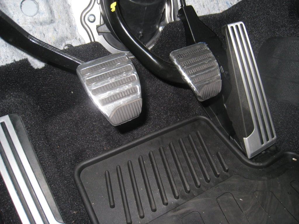 Three car pedals on a dark floor: clutch (left), brake (center), and accelerator (right).
