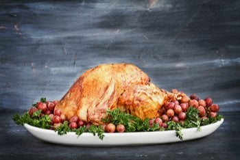 A Thanksgiving feast laid out on a wooden table, including roast turkey and a sliced holiday roast, alongside sides like corn, mushrooms, colorful tomatoes, and pumpkins.