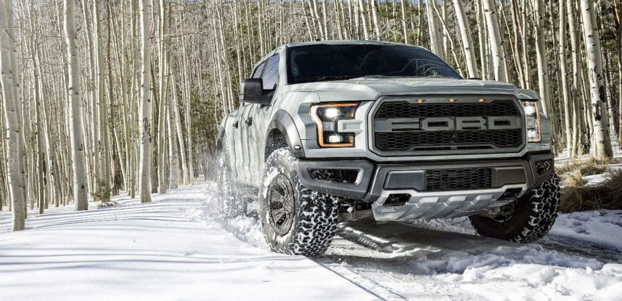 Dark gray Ford pickup truck with orange daytime running lights driving on a snowy mountain road.