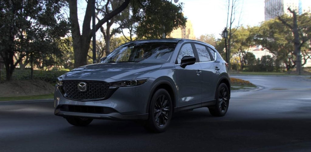 A dark gray Mazda CX-5 SUV parked on an asphalt road with trees and a building in the background