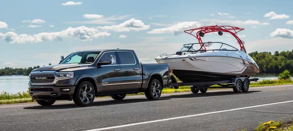 A dark gray truck tows a white boat on a trailer down a road beside a lake and trees under a cloudy sky.