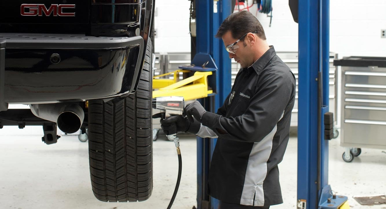 Mechanic in black coveralls using an impact wrench to change a tire on a lifted grey car.