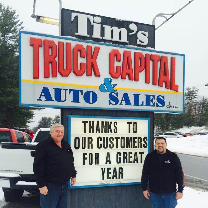 Two men stand beside a business sign for Tim's Truck Capital &amp; Auto Sales with a Thanks to our customers for a great year message.