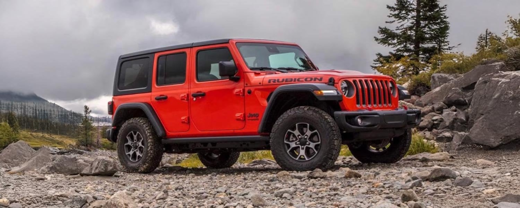 A Red Jeep Rubicon fords a rocky river, splashing water, with a forested mountain and snowy peaks in the background.