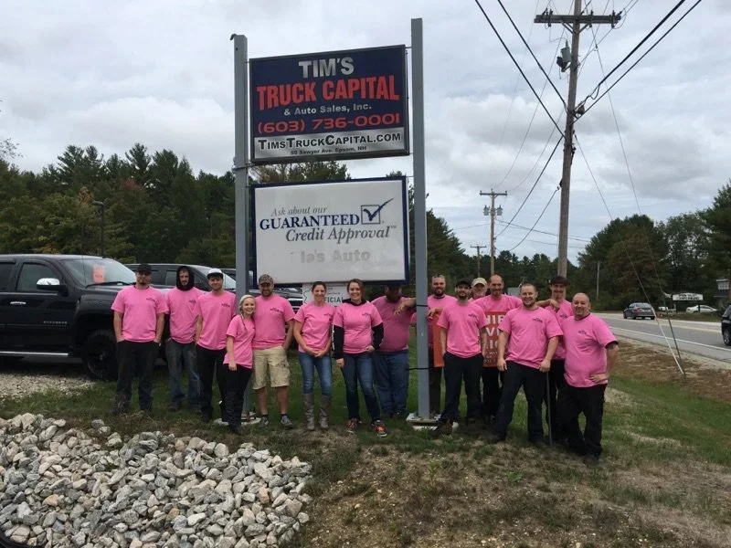 A group of staff in pink shirts stands in front of the Tim's Truck Capital sign and used trucks.
