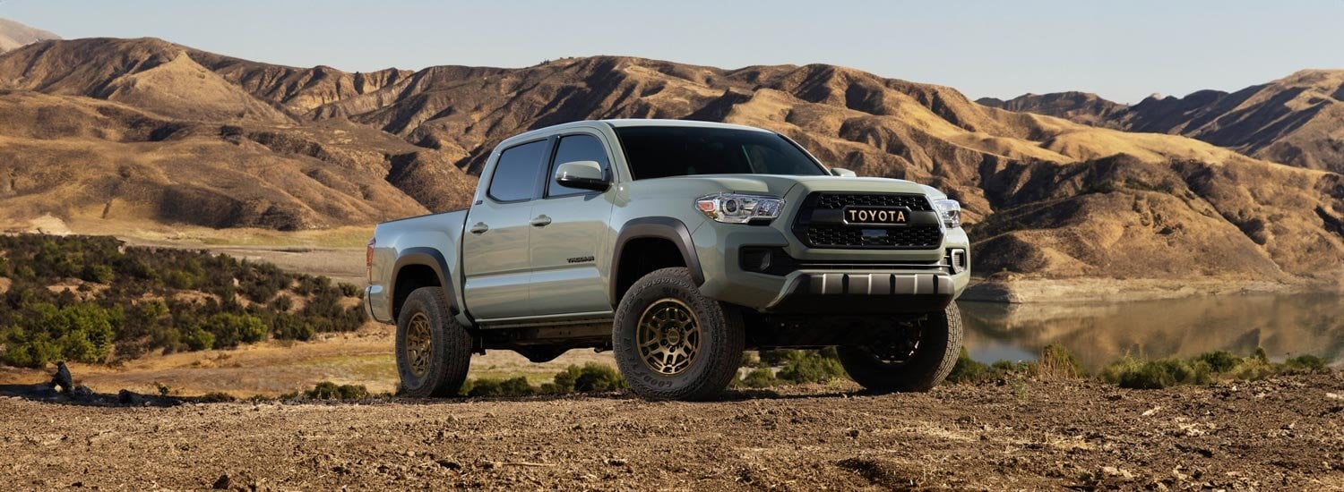 Light green Toyota pickup truck parked on a dirt hill overlooking a lake and rugged mountains.