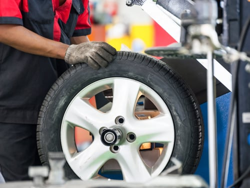 A young male mechanic in a gray overall and black gloves is carefully inspecting a car tire.