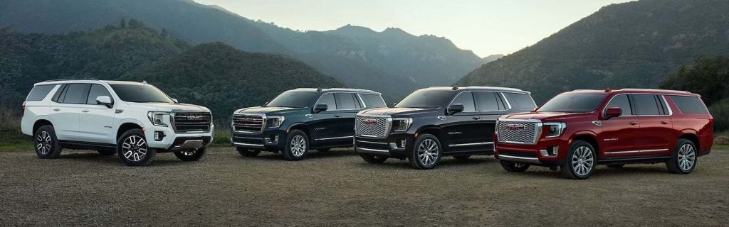 Four GMC Yukon SUVs in white, green, black, and red, lined up on a dirt road, with mountains in the background.